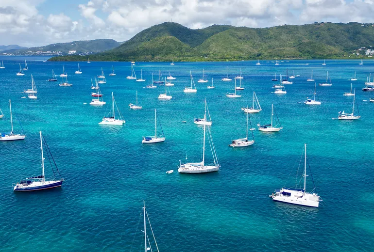 Aerial of a crowded sailboat anchorage in a turquoise bay with green hills