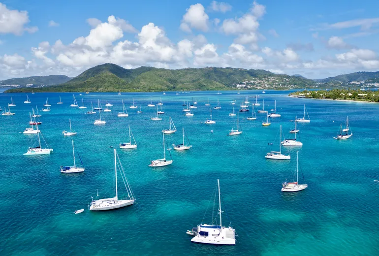 Aerial of an anchorage with a catamaran foreground and many sailboats off a green Caribbean coast