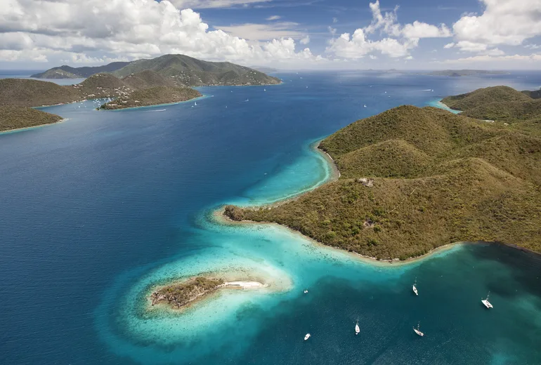 Aerial of green headlands, a reef-fringed cay and scattered anchored sailboats