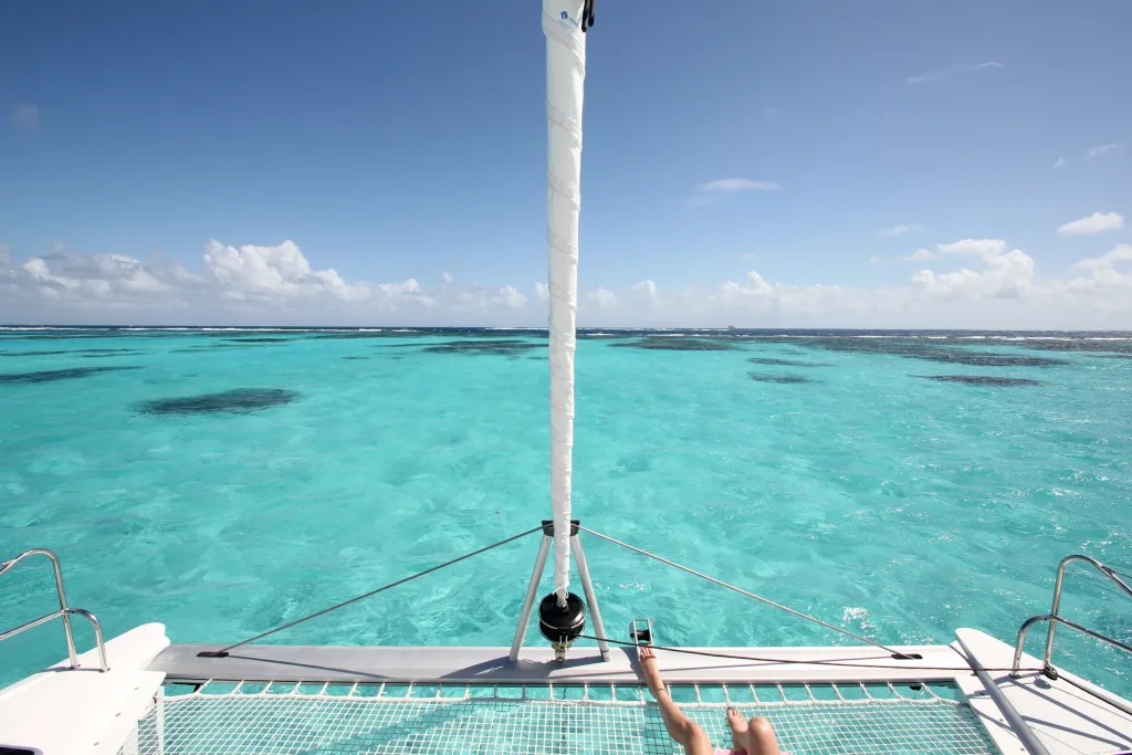 View from catamaran bow trampoline over turquoise reef-dappled shallows, person's leg visible