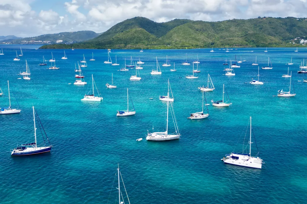 Aerial of crowded sailboat anchorage in turquoise bay with green hills behind