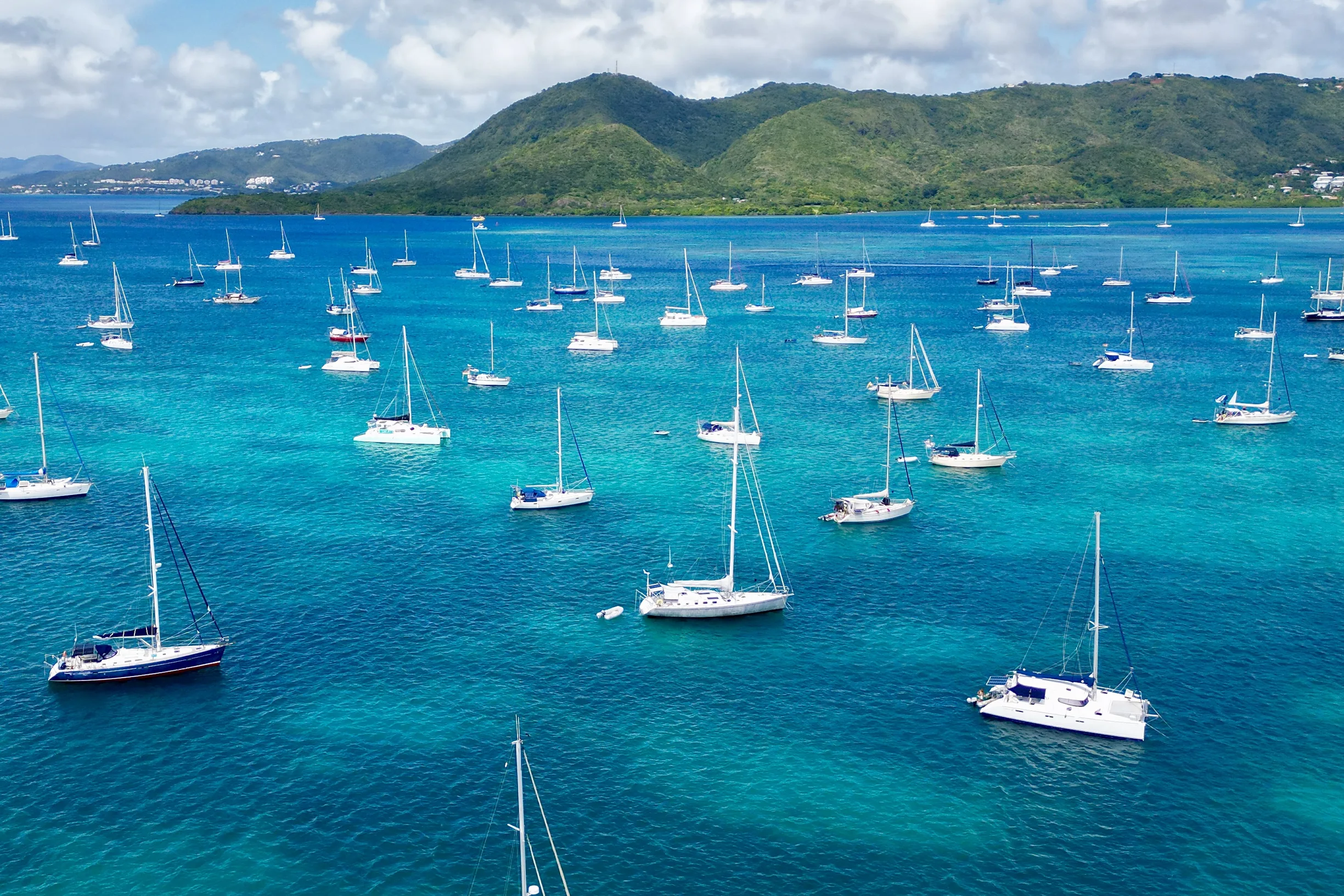 Aerial of crowded sailboat anchorage in turquoise bay with green hills behind
